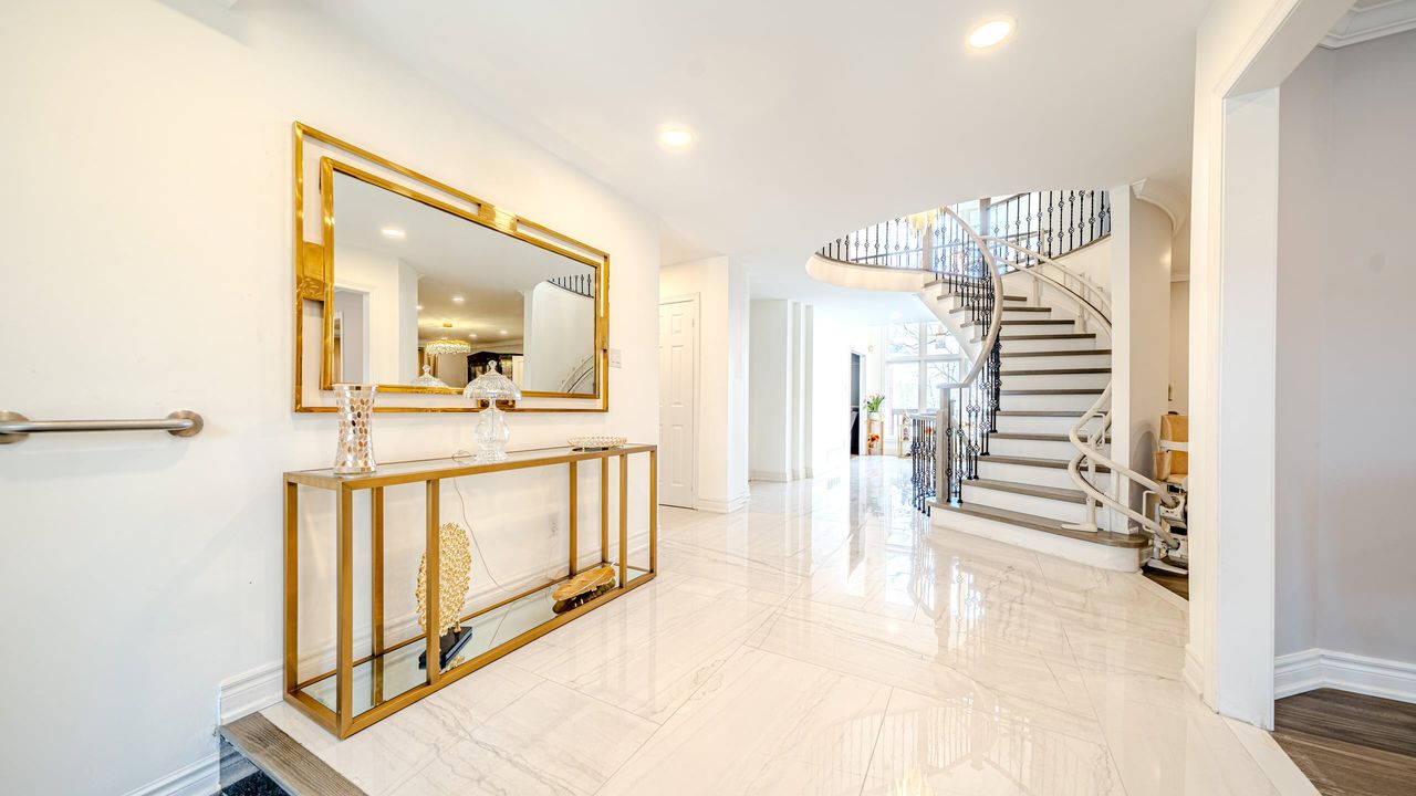 Entry hall with large glossy white veined floor tiles, a gold mirror over a console and a curved staircase
