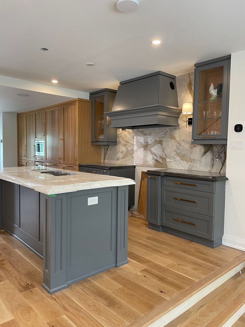 Grey shaker kitchen with a custom range hood, stone slab backsplash, brass pulls and oak floors