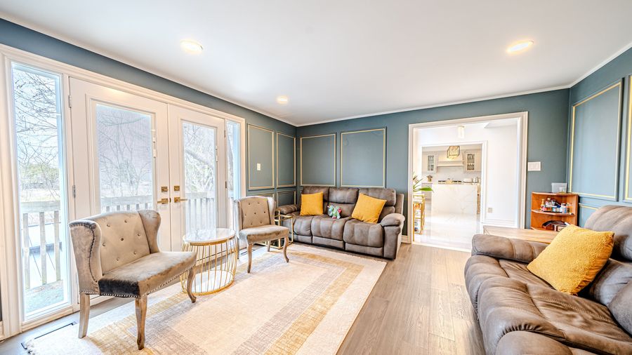 Living room with teal walls, gold trimmed box moulding, plank flooring and French doors, open to a white kitchen