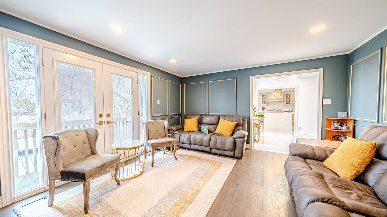 Living room with teal walls, gold trimmed box moulding, plank flooring and French doors, open to a white kitchen