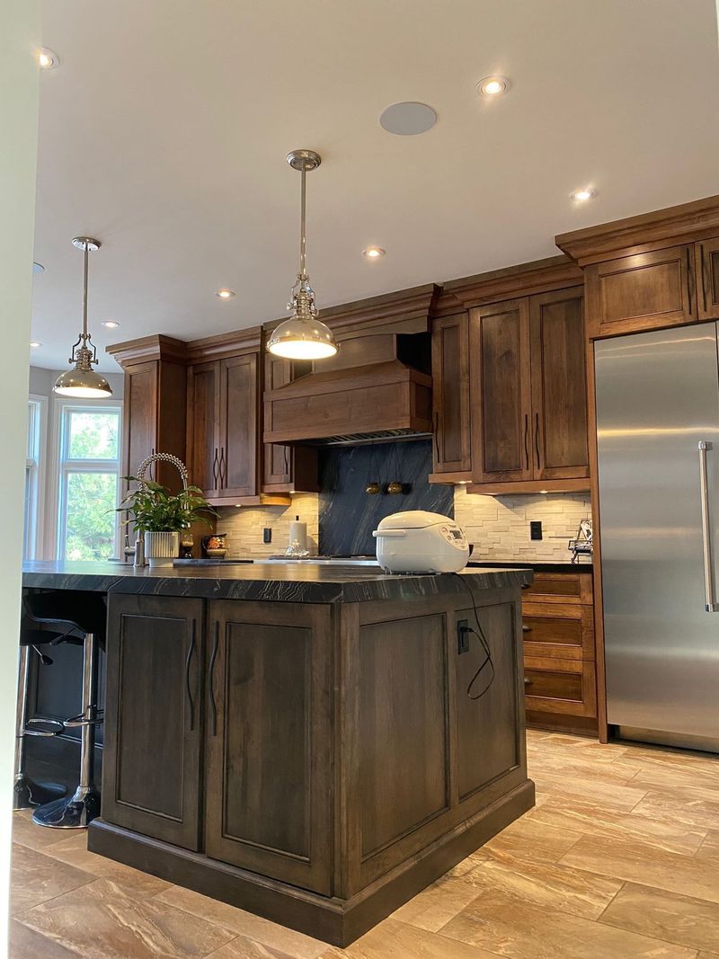 Brown stained kitchen with a black granite island, wood range hood and polished nickel pendants