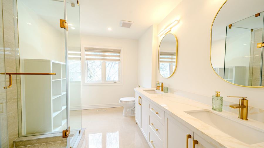 Bathroom with a glass shower, double vanity with veined countertop, gold faucets, oval mirrors and glossy tile floor