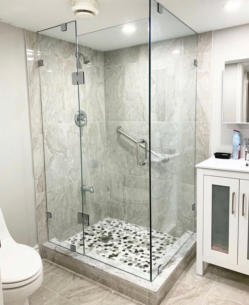 Glass corner shower with grey marble-look tile, pebble mosaic floor and a white vanity