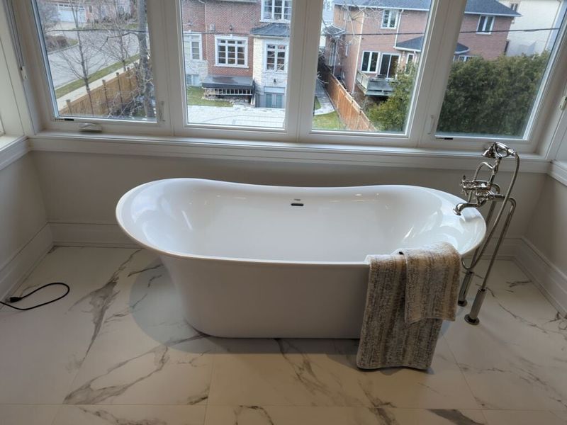 Freestanding soaker tub with a floor mounted tub filler under a wide window on marble look porcelain tile