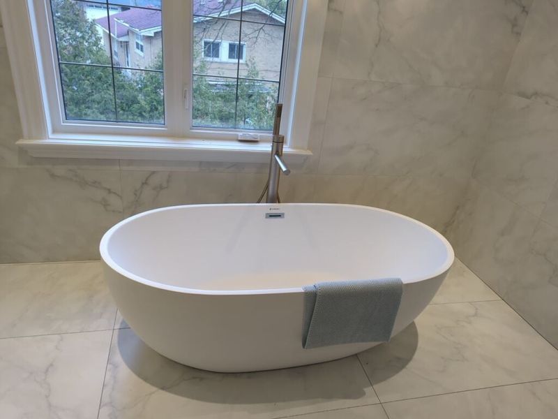 Freestanding oval soaker tub with a floor mounted filler beneath a window in marble look tile