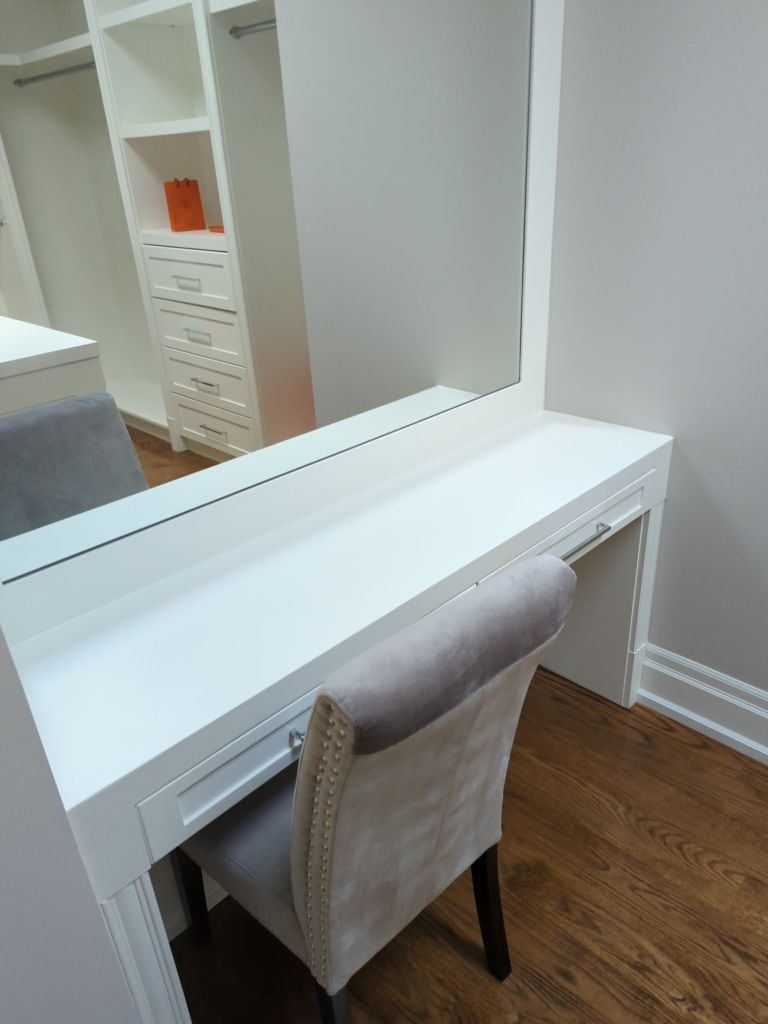 Built-in white makeup desk with a large mirror and an upholstered chair in a walk-in closet