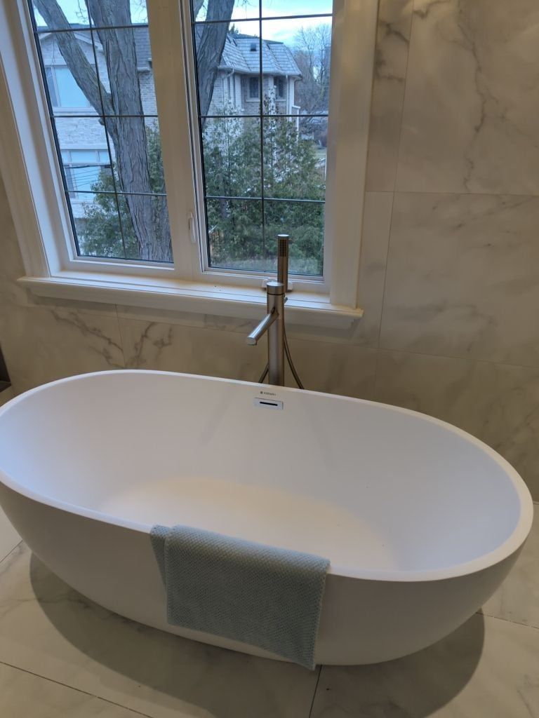 Freestanding oval soaker tub with a brushed floor mounted filler under a window in marble look tile