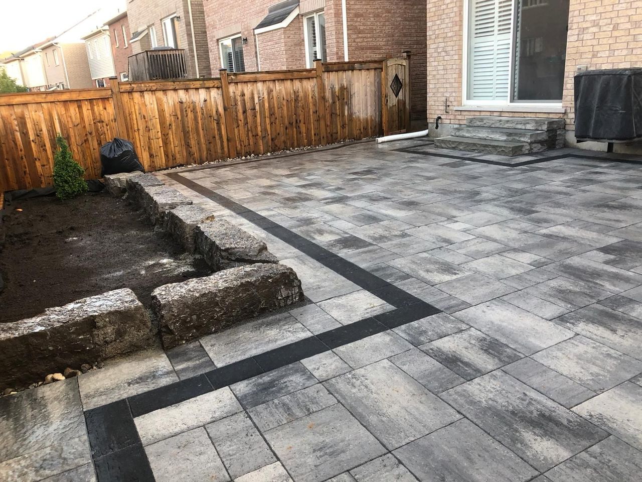 Backyard patio of grey interlocking pavers with charcoal border, armour stone garden wall and stone steps