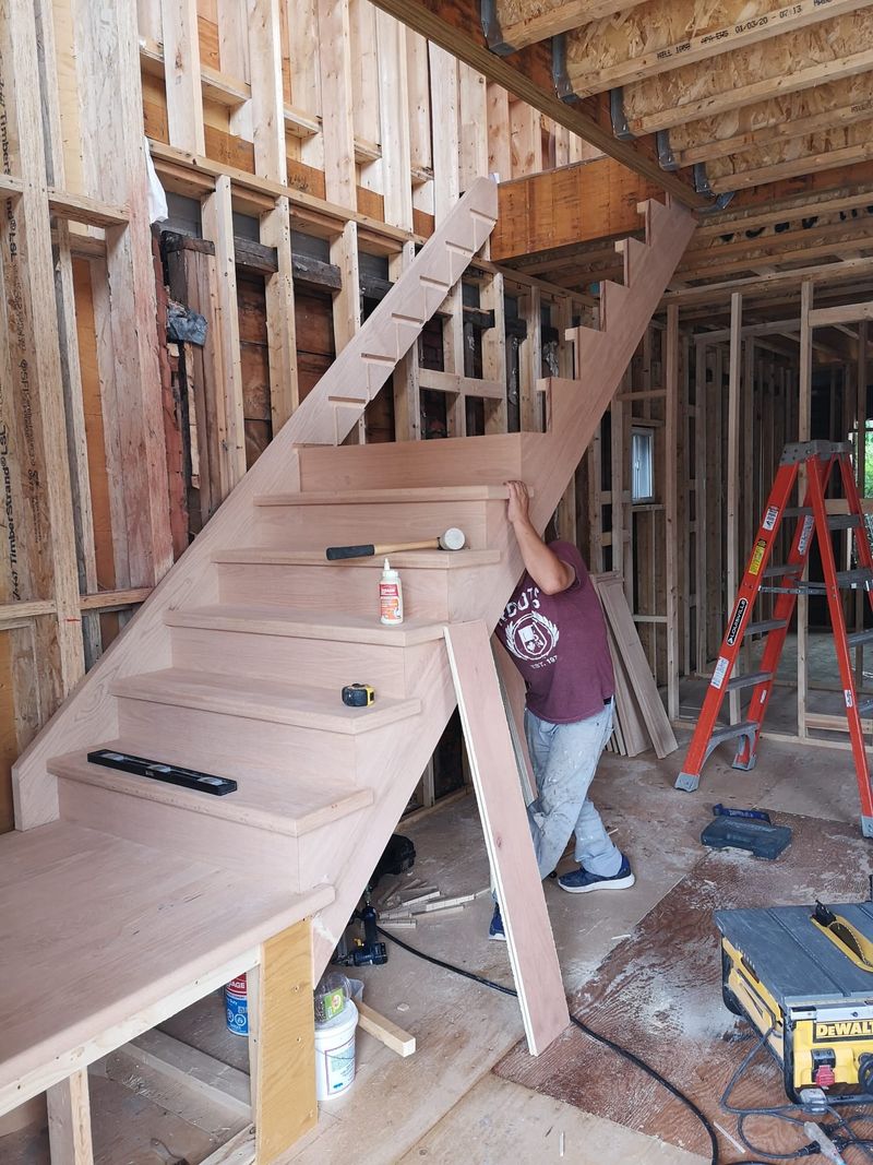 An oak staircase under construction inside a wood framed house, with a carpenter fitting the stringer