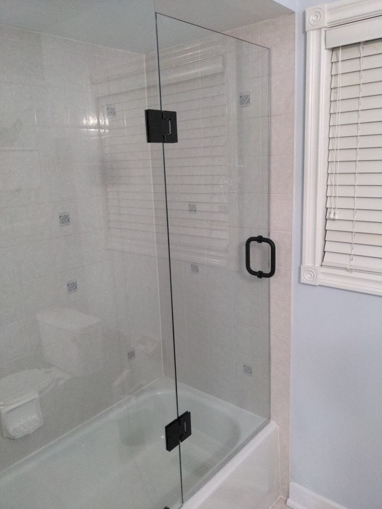 Hinged glass shower door with matte black hardware installed on a tiled tub