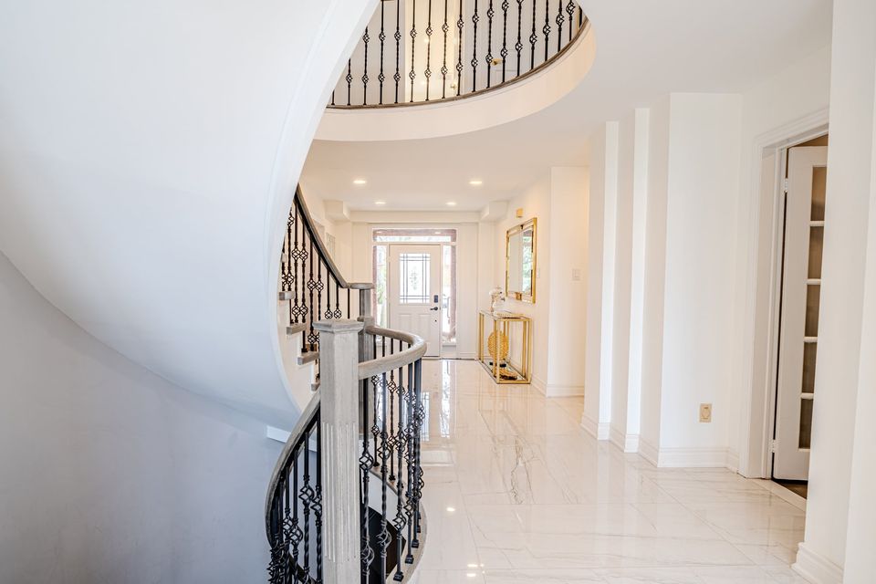 Curved staircase with black iron pickets and a grey stained handrail beside a polished white tile hallway with pot lights, leading to the front door